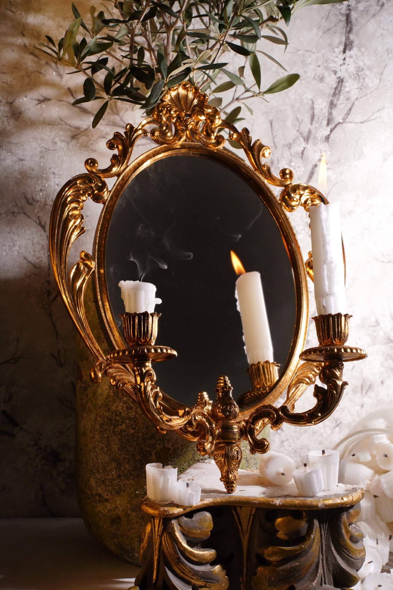 Dekoracyjne, złote lustro owalne z palącymi się świecami w świecznikach, odbijające płomień i dym, na tle marmurkowej ściany i gałązek oliwnych, sfotografowane w Krakowie.
