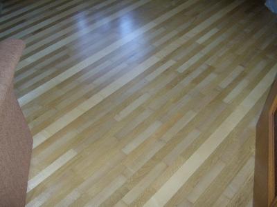 Ukośne ułożenie jasnych, lakierowanych desek parkietowych, widoczne delikatne refleksy światła, fragment brązowego mebla w rogu kadru.