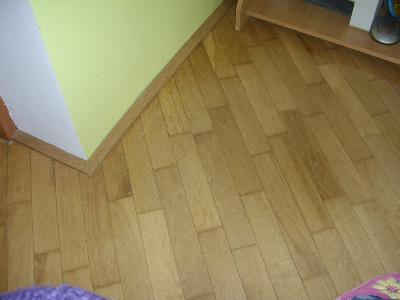 Jasny parkiet ułożony ukośnie względem ściany pomalowanej na biało i jasnozielono, widoczna listwa przypodłogowa i fragment półki z książkami.