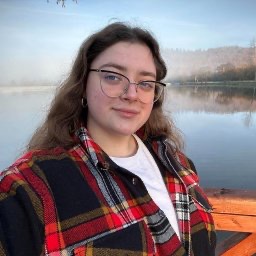 Portret kobiety w okularach i flanelowej koszuli na tle jeziora i drzew. Uśmiechnięta twarz, naturalne światło, delikatna mgła nad wodą. Widok na krajobraz.