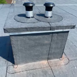 Szczegółowe ujęcie płaskiego dachu pokrytego papą z dwoma kominkami wentylacyjnymi, widoczne łączenia papy i uszczelnienia wokół kominków.