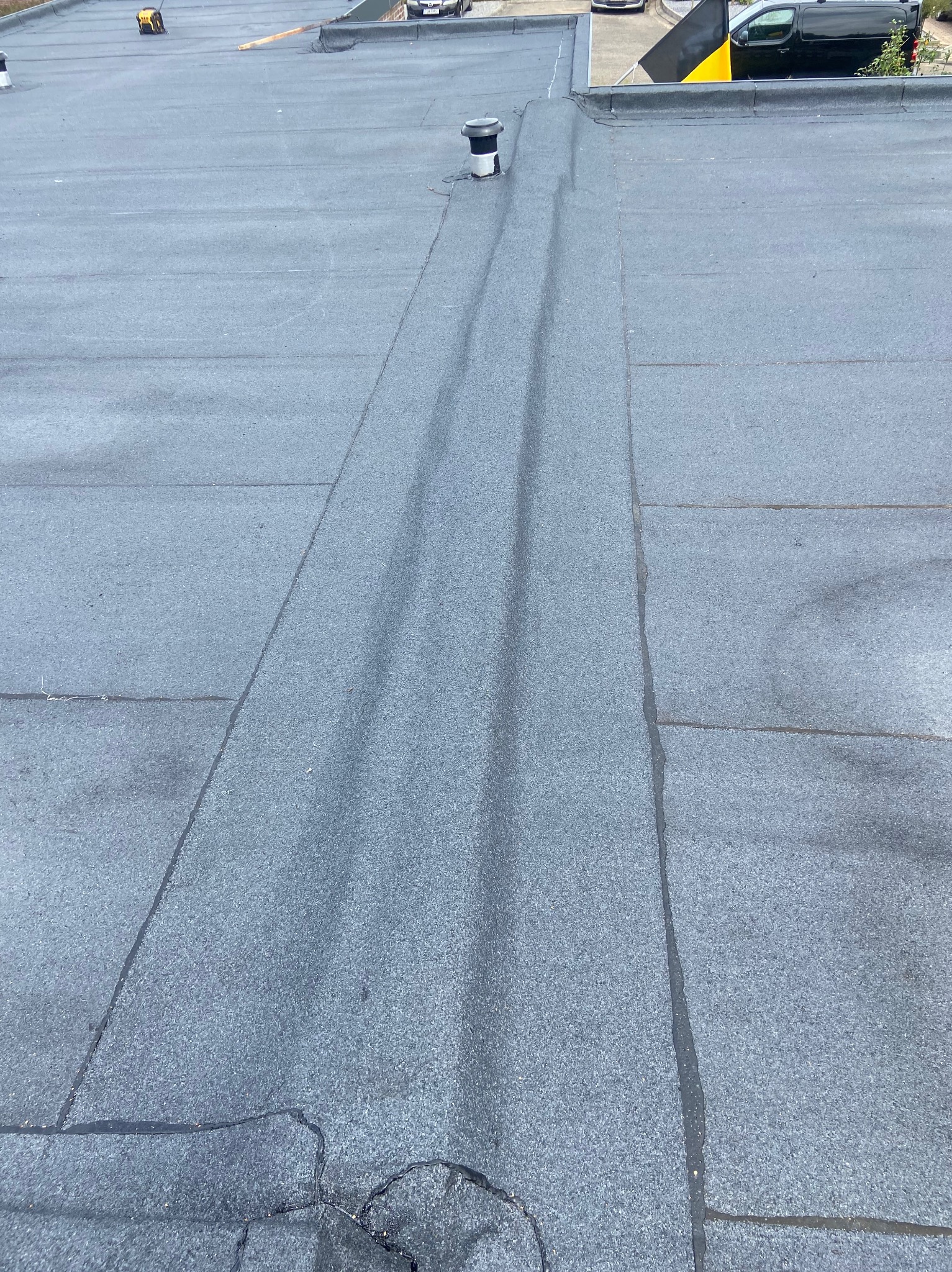 Płaski dach pokryty papą termozgrzewalną z widocznymi łączeniami i kominkiem wentylacyjnym; w tle zaparkowane samochody.