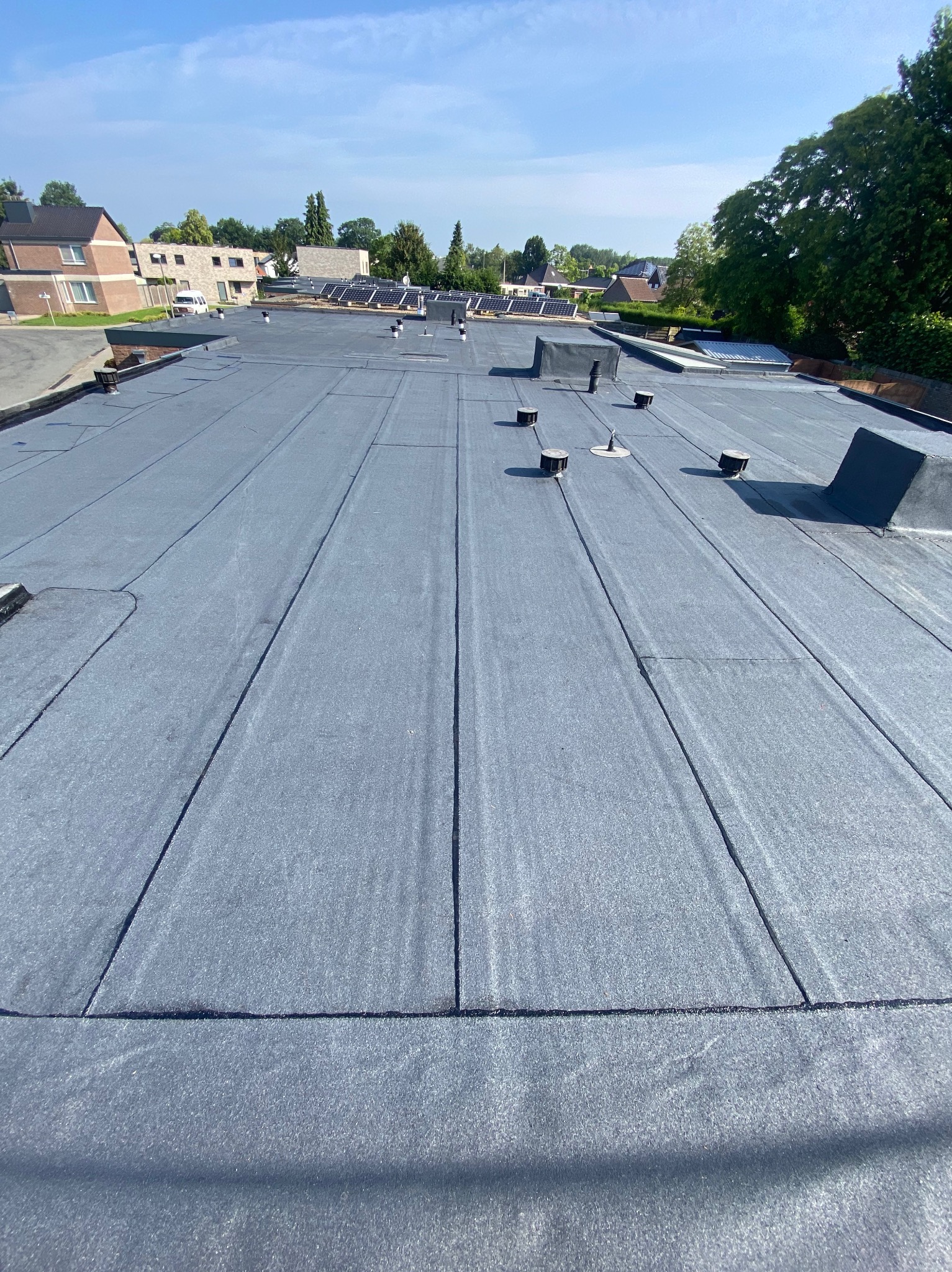 Płaski, szary dach pokryty papą termozgrzewalną z widocznymi odpowietrznikami i kominem wentylacyjnym, na tle zabudowań mieszkalnych w słoneczny dzień.