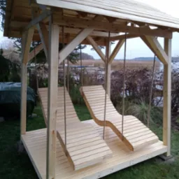 Drewniana altana z dwoma wiszącymi leżakami na linach, widok na jezioro w tle.