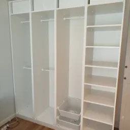 Biała, modułowa szafa garderobiana z półkami i drążkami, uzupełniona o kosze i tekstylne pojemniki, ustawiona na drewnianej podłodze, widok z przodu.