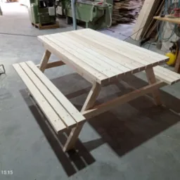 Nowy, jasny stół piknikowy z ławkami, wykonany z surowego drewna, ustawiony w warsztacie. Widoczne narzędzia i materiały stolarskie w tle.