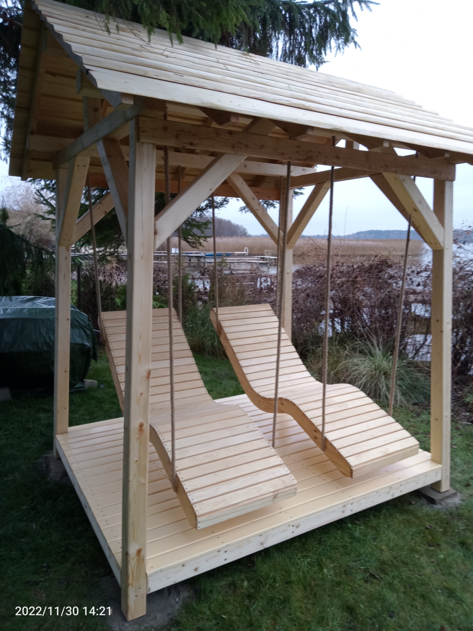 Drewniana altana z dwoma wiszącymi leżakami na linach, widok na jezioro w tle.