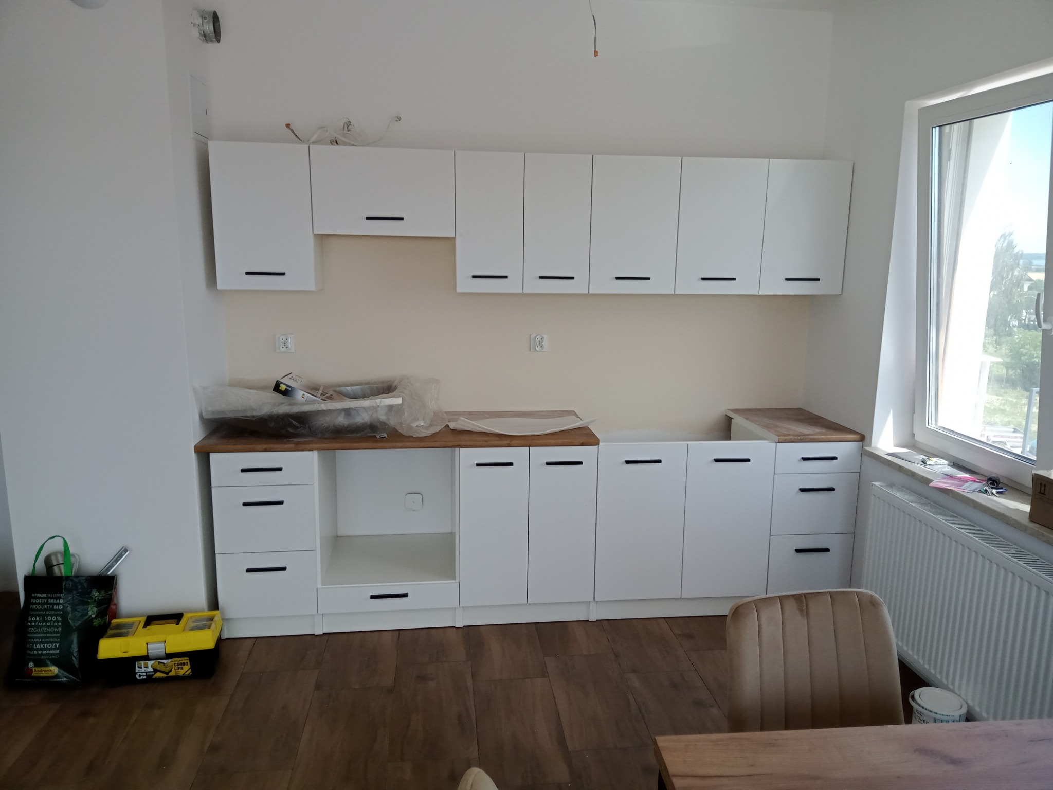 Wnętrze kuchni w trakcie urządzania: białe szafki z drewnianymi blatami, narzędzia w skrzynce obok, częściowo rozpakowane elementy, widok na okno.