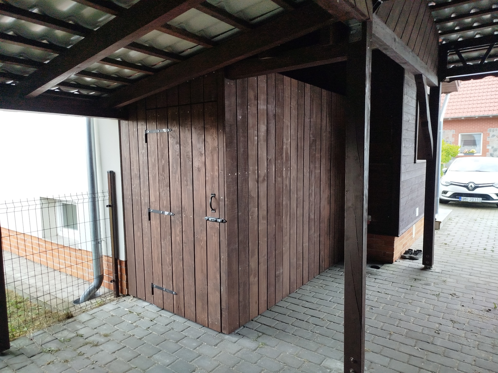 Zadaszona, drewniana konstrukcja z drzwiami, przypominająca schowek lub małą altanę, na posesji z kostki brukowej i widocznym budynkiem w tle. Ciemne drewno, solidne wykonanie.