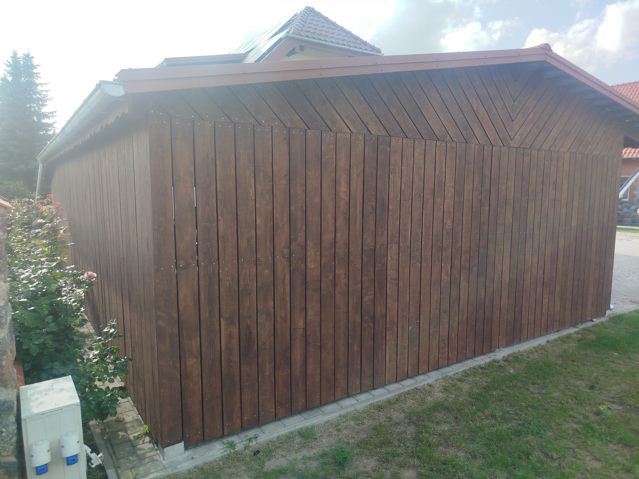 Ciemnobrązowa, drewniana konstrukcja z pionowych desek, zwieńczona dachem, widoczna z boku, otoczona zielenią. Na górze widoczny fragment dachu budynku.