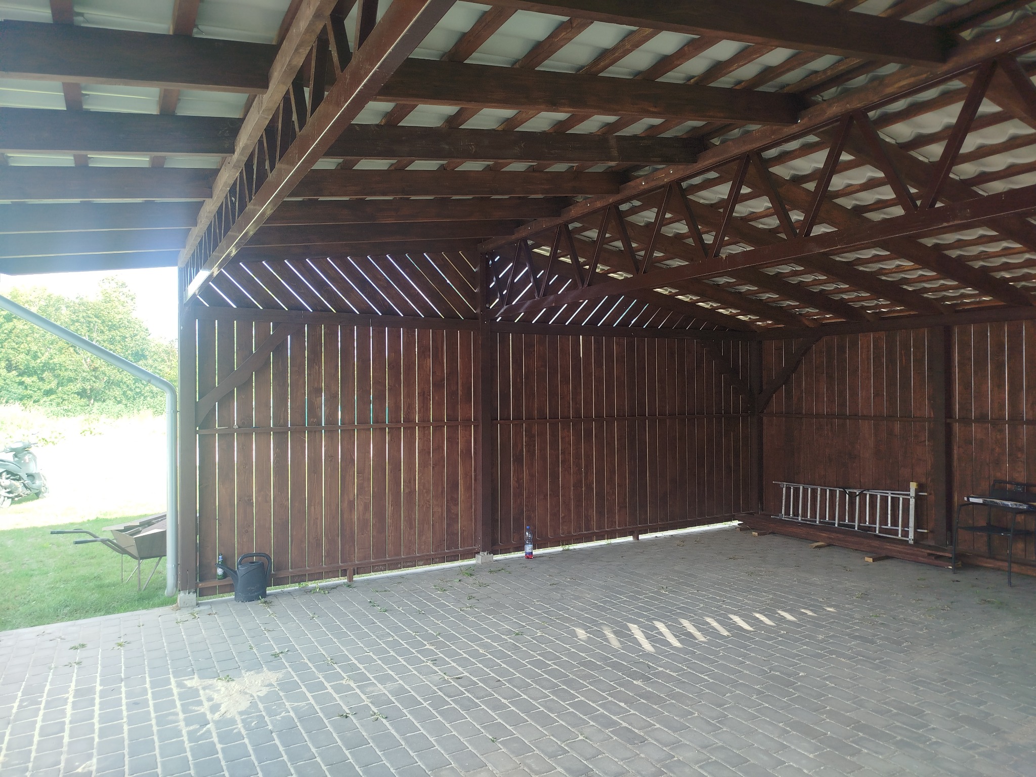 Zadaszona, drewniana konstrukcja z widoczną więźbą dachową i ażurowymi ścianami. Kostka brukowa na posadzce. Na zewnątrz zieleń i sprzęt ogrodowy.