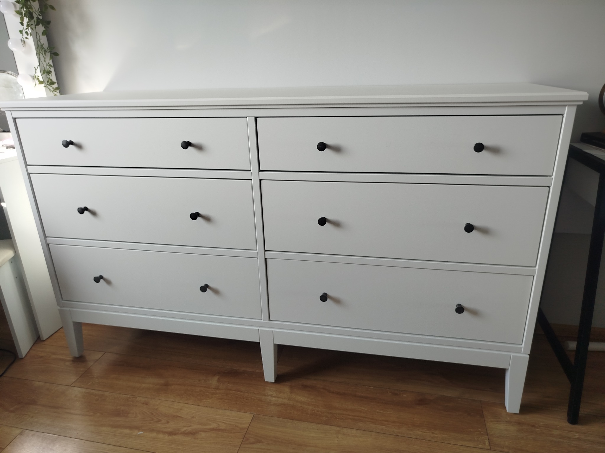 Biała komoda z sześcioma szufladami na drewnianej podłodze, z czarnymi okrągłymi uchwytami, sfotografowana z przodu, z widocznym fragmentem blatu biurka po prawej stronie.