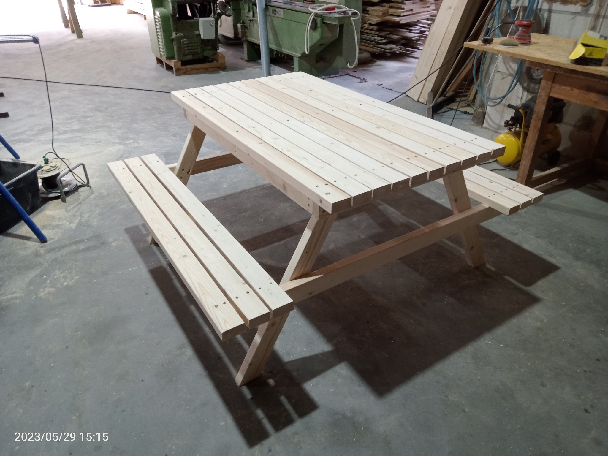 Nowy, jasny stół piknikowy z ławkami, wykonany z surowego drewna, ustawiony w warsztacie. Widoczne narzędzia i materiały stolarskie w tle.