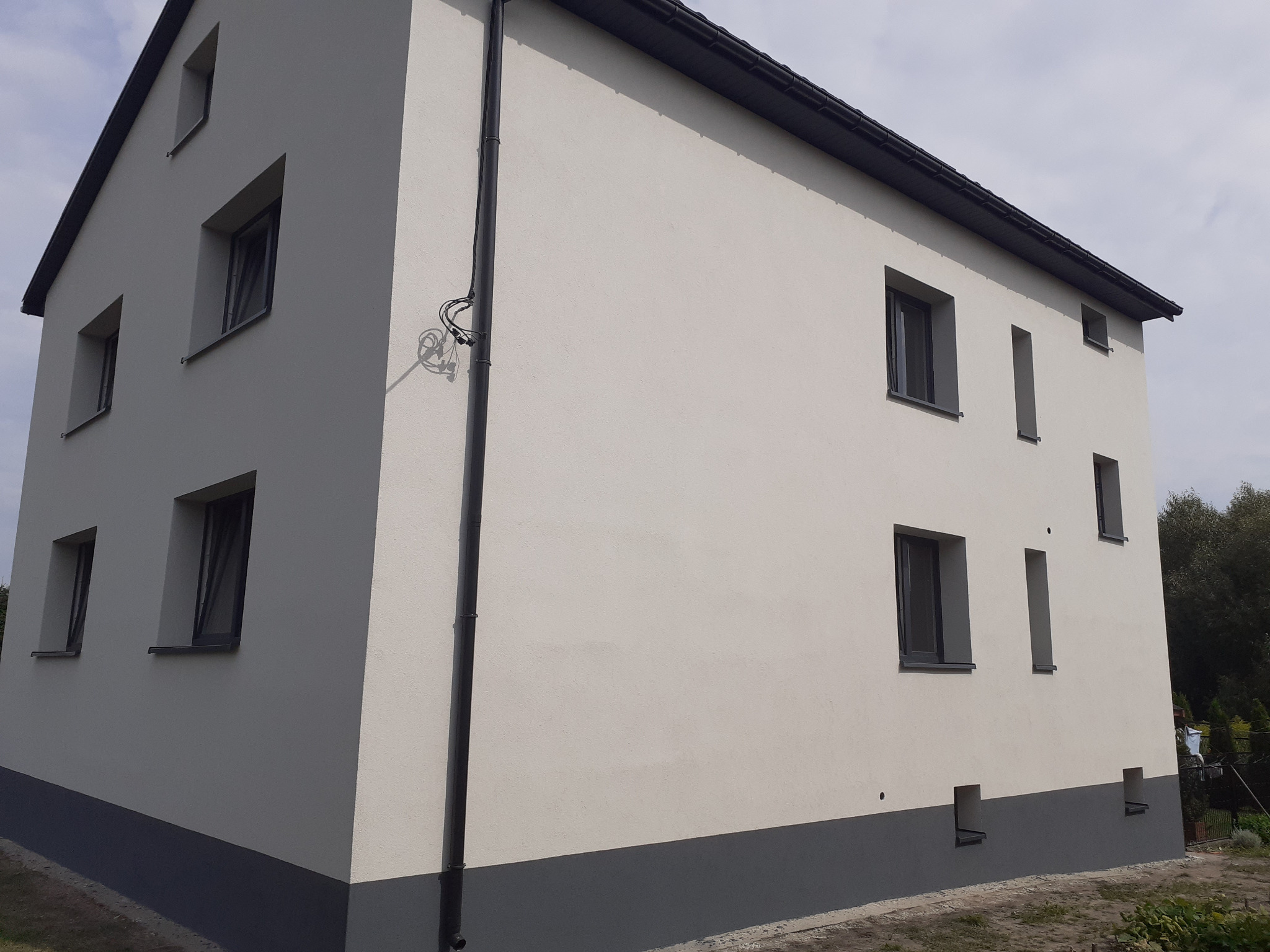 Dwukondygnacyjny dom z jasną elewacją i ciemnoszarą podmurówką, widok z boku na narożnik budynku z widoczną instalacją elektryczną na ścianie.