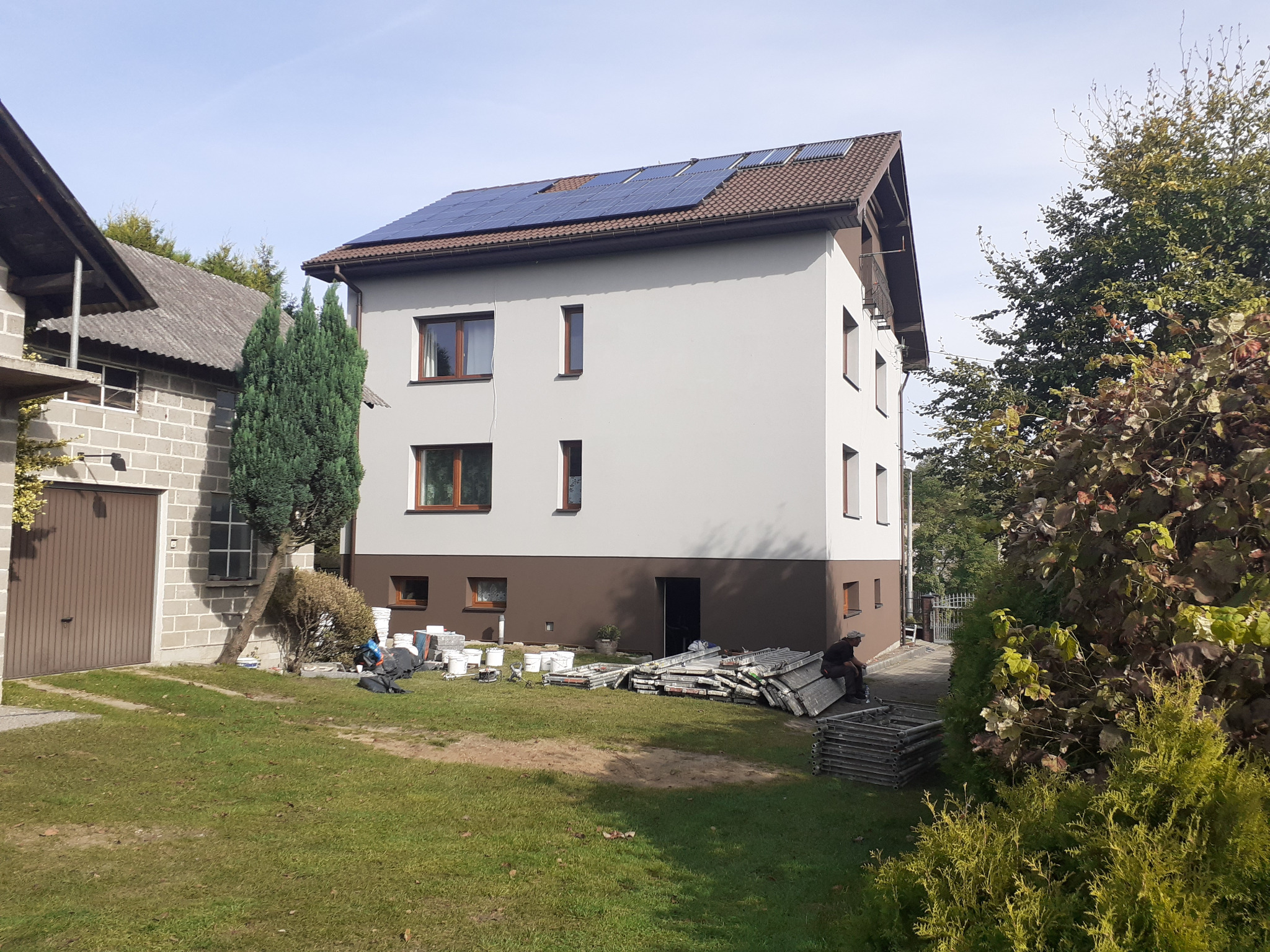 Dwukondygnacyjny dom po remoncie elewacji z brązowym dołem i białymi ścianami, brązowymi ramami okiennymi, panelami słonecznymi na dachu, widoczne rusztowania i pracownicy na trawniku.