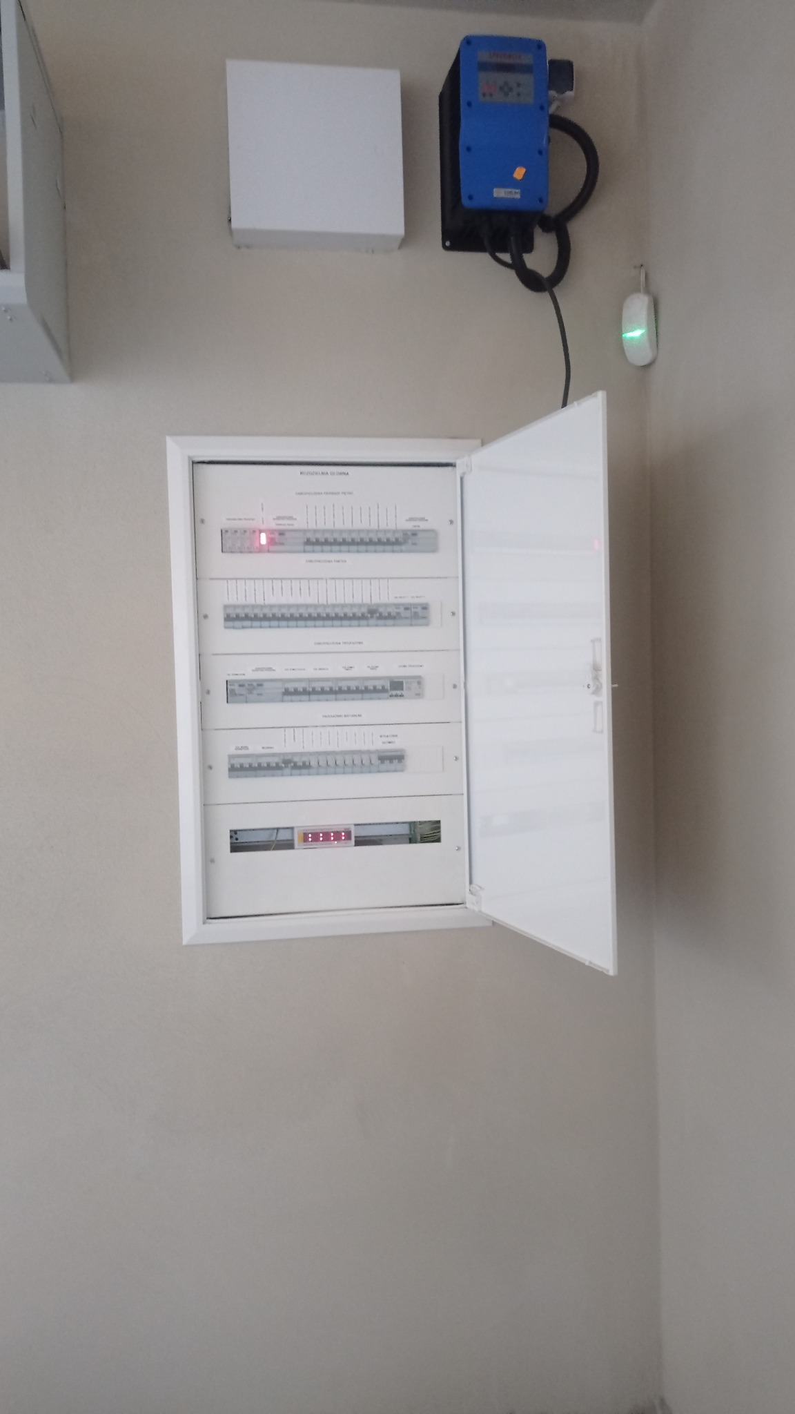 Rozdzielnica elektryczna z otwartymi drzwiczkami, widoczne zabezpieczenia i moduły. Na ścianie dodatkowo licznik energii elektrycznej i czujnik.