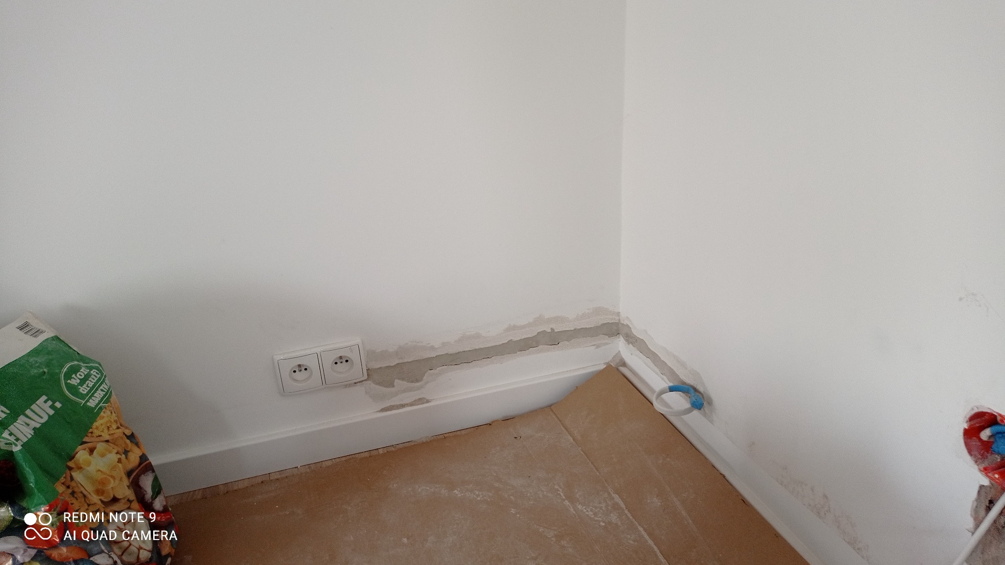 Biała ściana z gniazdkiem elektrycznym, widoczne ślady po kuciu i instalacji. Częściowo zakryte podłogi. Przygotowanie do prac wykończeniowych.