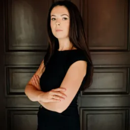 Portret kobiety w czarnej sukience, z założonymi rękami, na tle ciemnych drzwi w Częstochowie. Kobieta ma długie, ciemne włosy i nosi pierścionek.