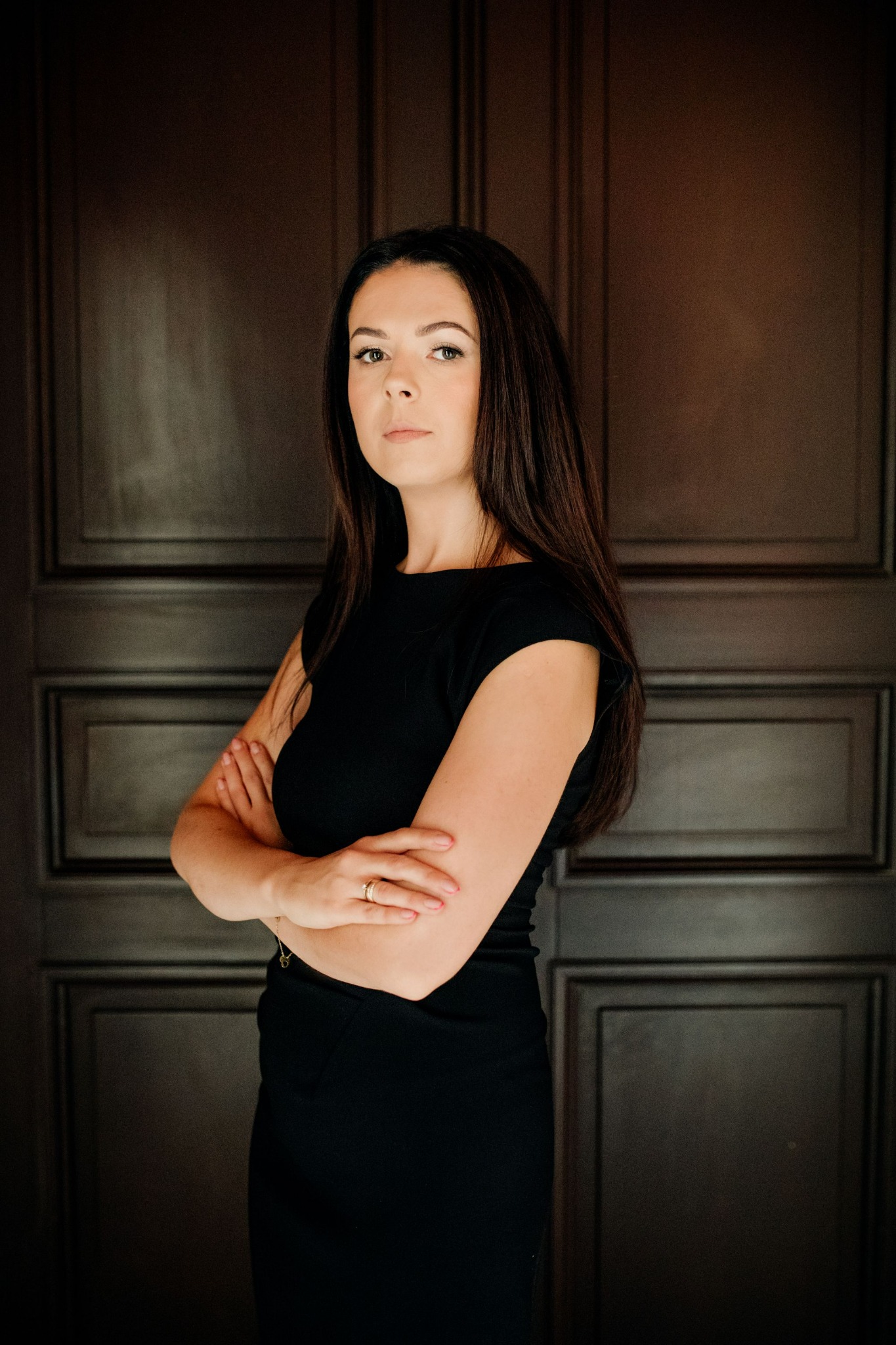 Portret kobiety w czarnej sukience, z założonymi rękami, na tle ciemnych drzwi w Częstochowie. Kobieta ma długie, ciemne włosy i nosi pierścionek.