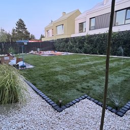 SZEF OGRODU - Nowoczesny ogród z trawnikiem, systemem nawadniania i dekoracyjnym obrzeżem z kamyków. W tle budynki o minimalistycznej architekturze i przygotowane miejsce pod budowę.