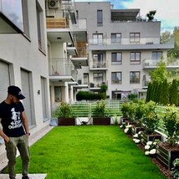 SZEF OGRODU - Zadbany trawnik przed nowoczesnym budynkiem mieszkalnym z białymi kwiatami w metalowych donicach. Mężczyzna w czarnej koszulce stoi z boku, obserwując ogród.