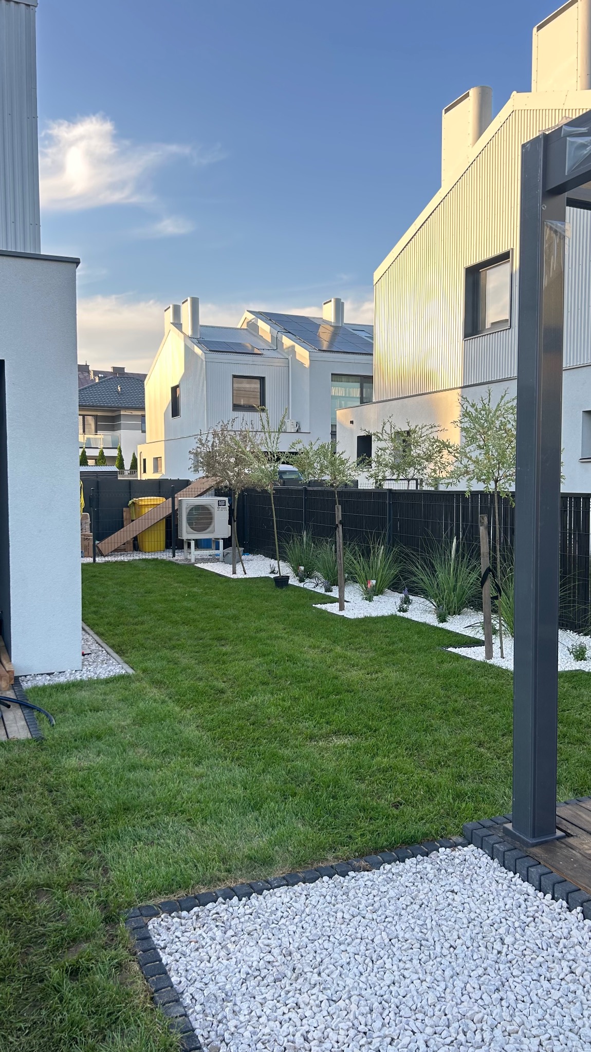 Nowoczesny ogród z trawnikiem, białymi kamieniami i młodymi drzewkami wzdłuż czarnego ogrodzenia na tle białych domów z panelami słonecznymi.