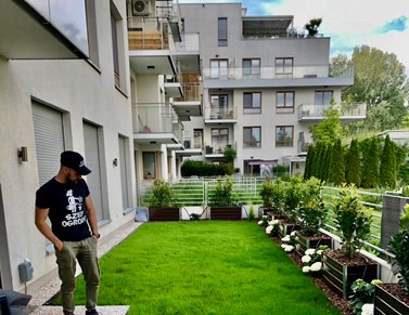 Zadbany trawnik przed nowoczesnym budynkiem mieszkalnym z białymi kwiatami w metalowych donicach. Mężczyzna w czarnej koszulce stoi z boku, obserwując ogród.