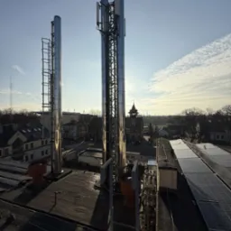 Błyszczące, metalowe kominy wentylacyjne z drabinkami i antena telekomunikacyjna na dachu budynku usługowego z panelami słonecznymi w słoneczny dzień.