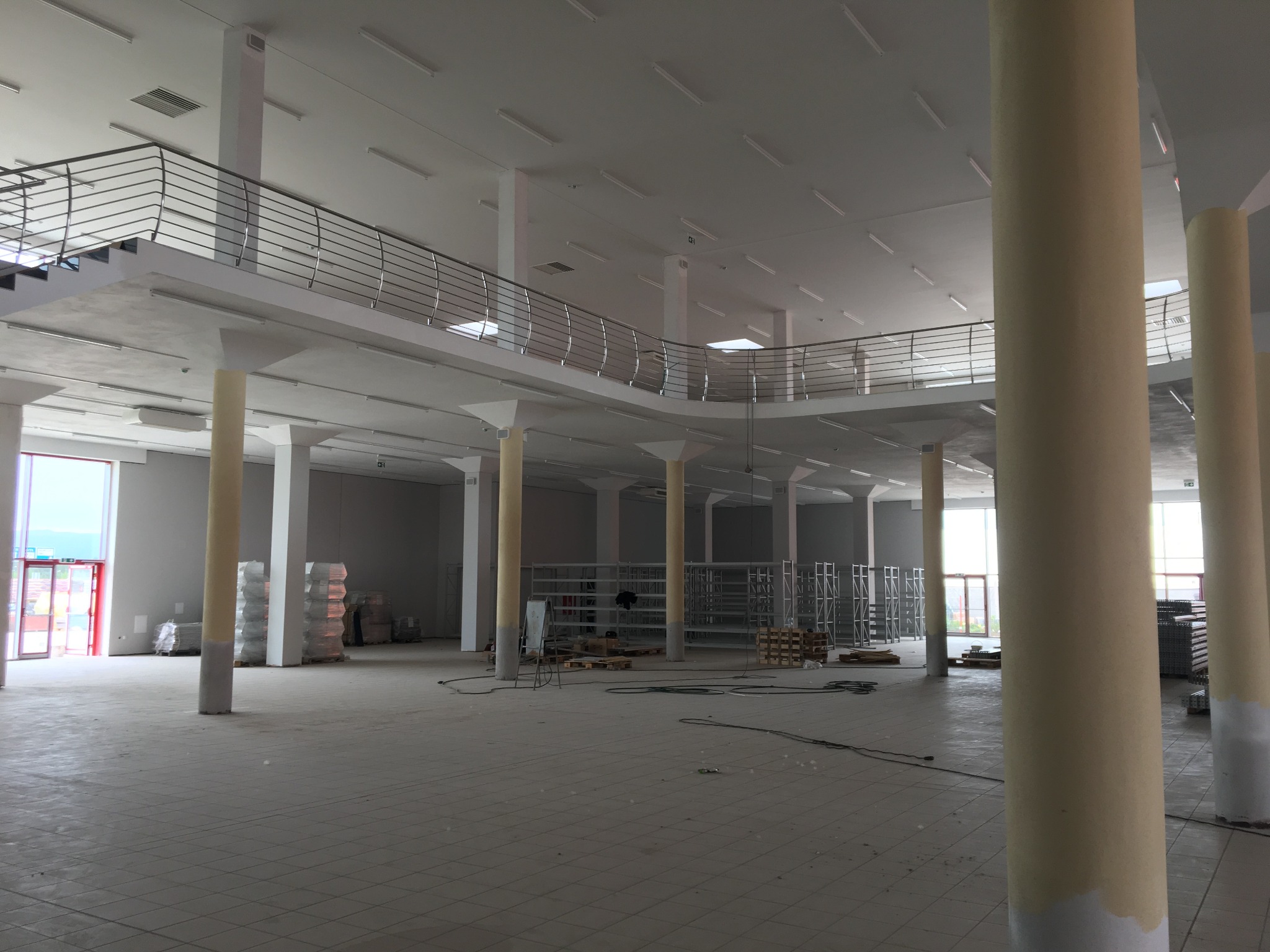 Wnętrze hali usługowej w trakcie wykańczania, widoczne kolumny, antresola z metalową balustradą, regały i materiały budowlane.