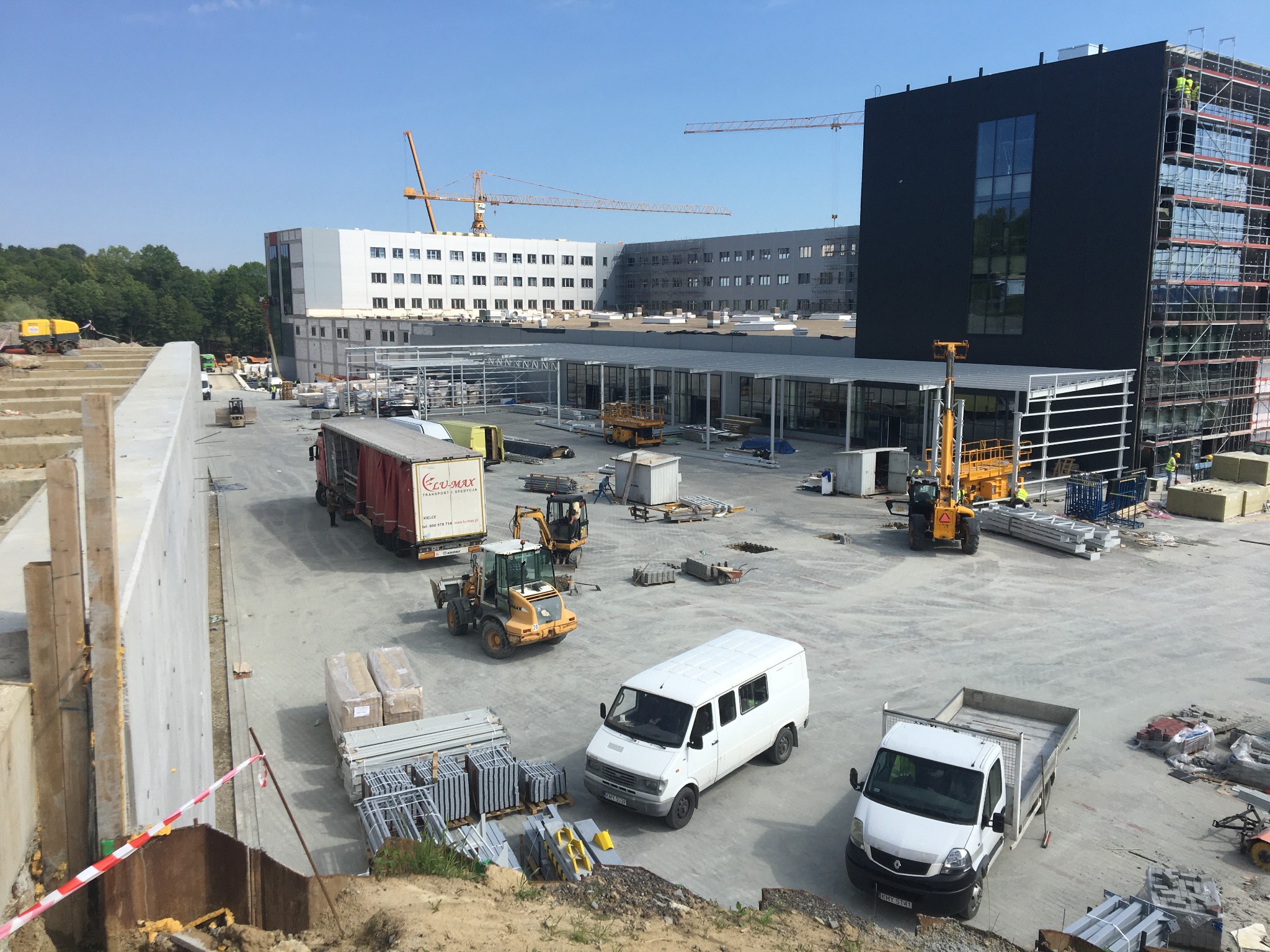 Plac budowy z widocznymi elementami konstrukcji stalowych, maszynami budowlanymi i dwoma budynkami w tle, jeden z rusztowaniem.