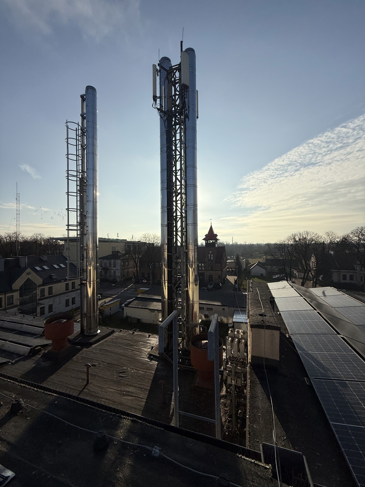 Błyszczące, metalowe kominy wentylacyjne z drabinkami i antena telekomunikacyjna na dachu budynku usługowego z panelami słonecznymi w słoneczny dzień.