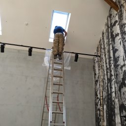 RTD TECHNOLOGY Robert Liberek - Pracownik na drabinie montuje oświetlenie sufitowe typu reflektor szynowy, zabezpieczone folią, w pomieszczeniu z drewnianymi belkami stropowymi i tapetą imitującą korę drzew.