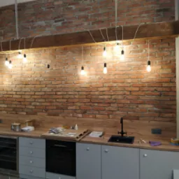 Kuchnia z ceglaną ścianą i wiszącymi żarówkami Edison na drewnianej belce, widoczne gniazdka elektryczne i blat z akcesoriami montażowymi.
