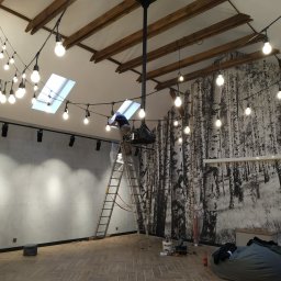 RTD TECHNOLOGY Robert Liberek - Pracownik na drabinie instaluje girlandę żarówkową w pomieszczeniu z betonową ścianą i fototapetą z motywem brzozowego lasu, widoczne narzędzia i elementy oświetlenia.