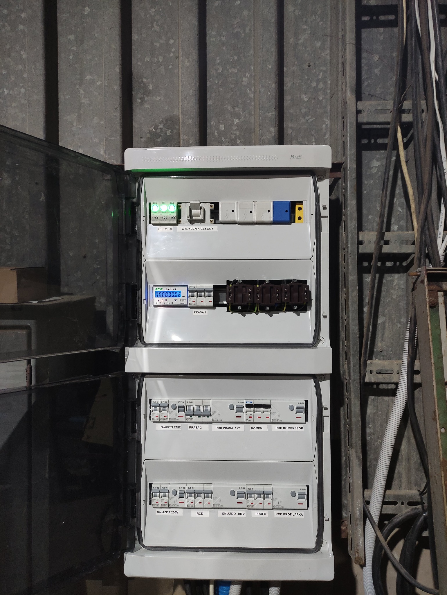 Rozdzielnica elektryczna z otwartymi drzwiczkami, widoczne zabezpieczenia, bezpieczniki i licznik energii elektrycznej. Na górze listwa z sygnalizacją świetlną i opisem 'Wyłącznik główny'.