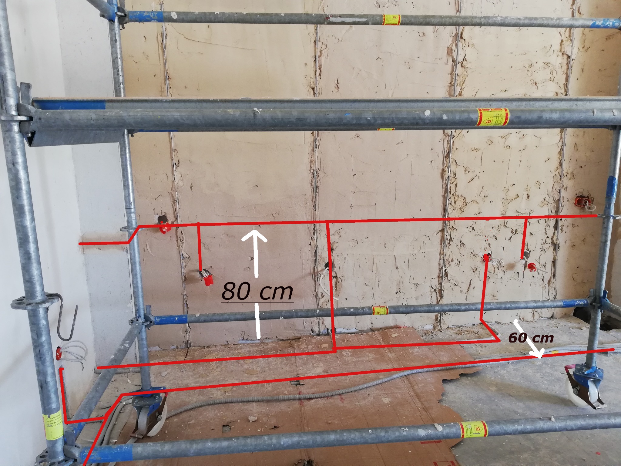 Szkielet rusztowania na tle ściany w trakcie remontu z zaznaczonymi na czerwono planowanymi trasami przewodów elektrycznych i podanymi odległościami 80 cm i 60 cm.