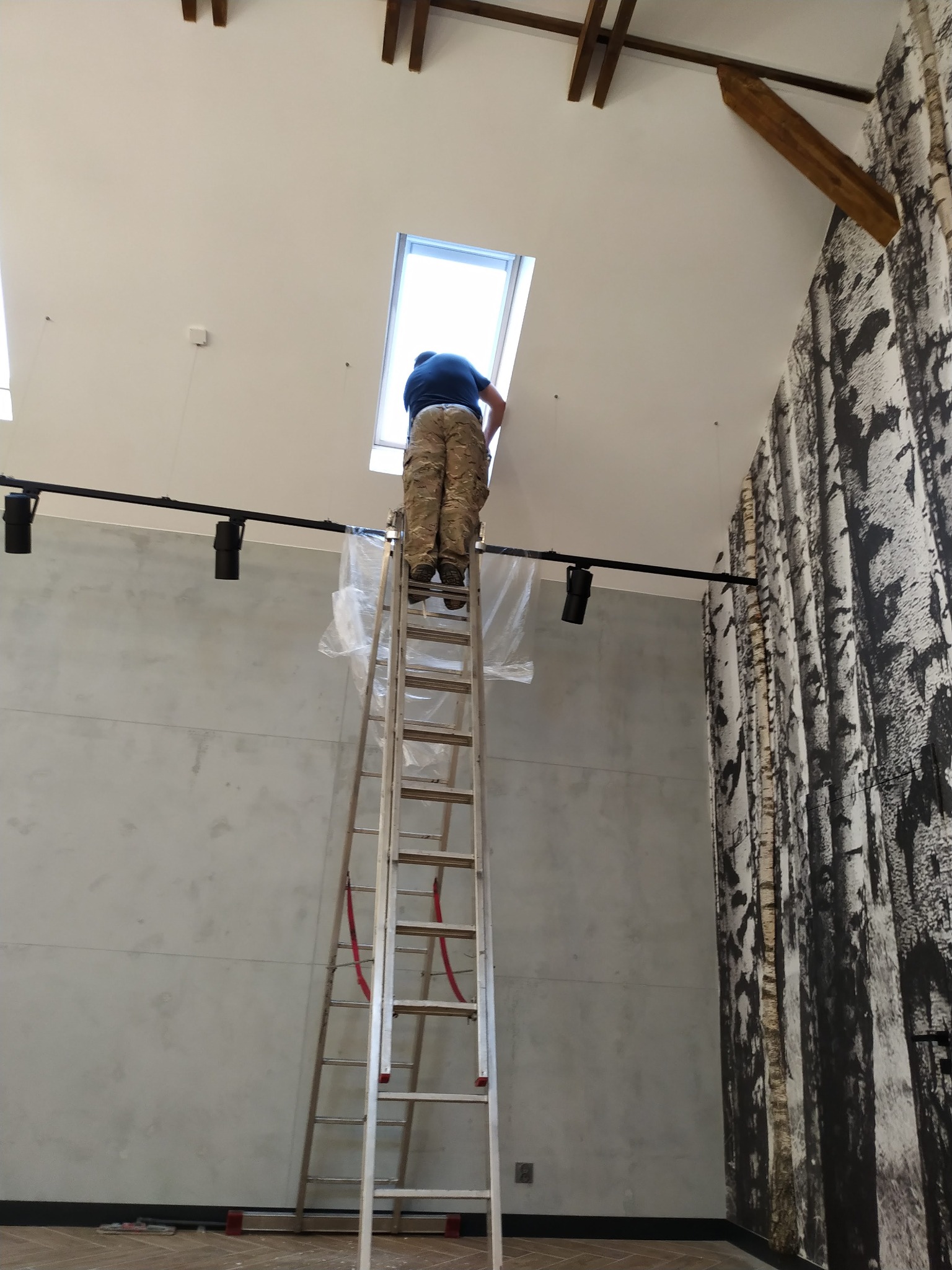 Pracownik na drabinie montuje oświetlenie sufitowe typu reflektor szynowy, zabezpieczone folią, w pomieszczeniu z drewnianymi belkami stropowymi i tapetą imitującą korę drzew.