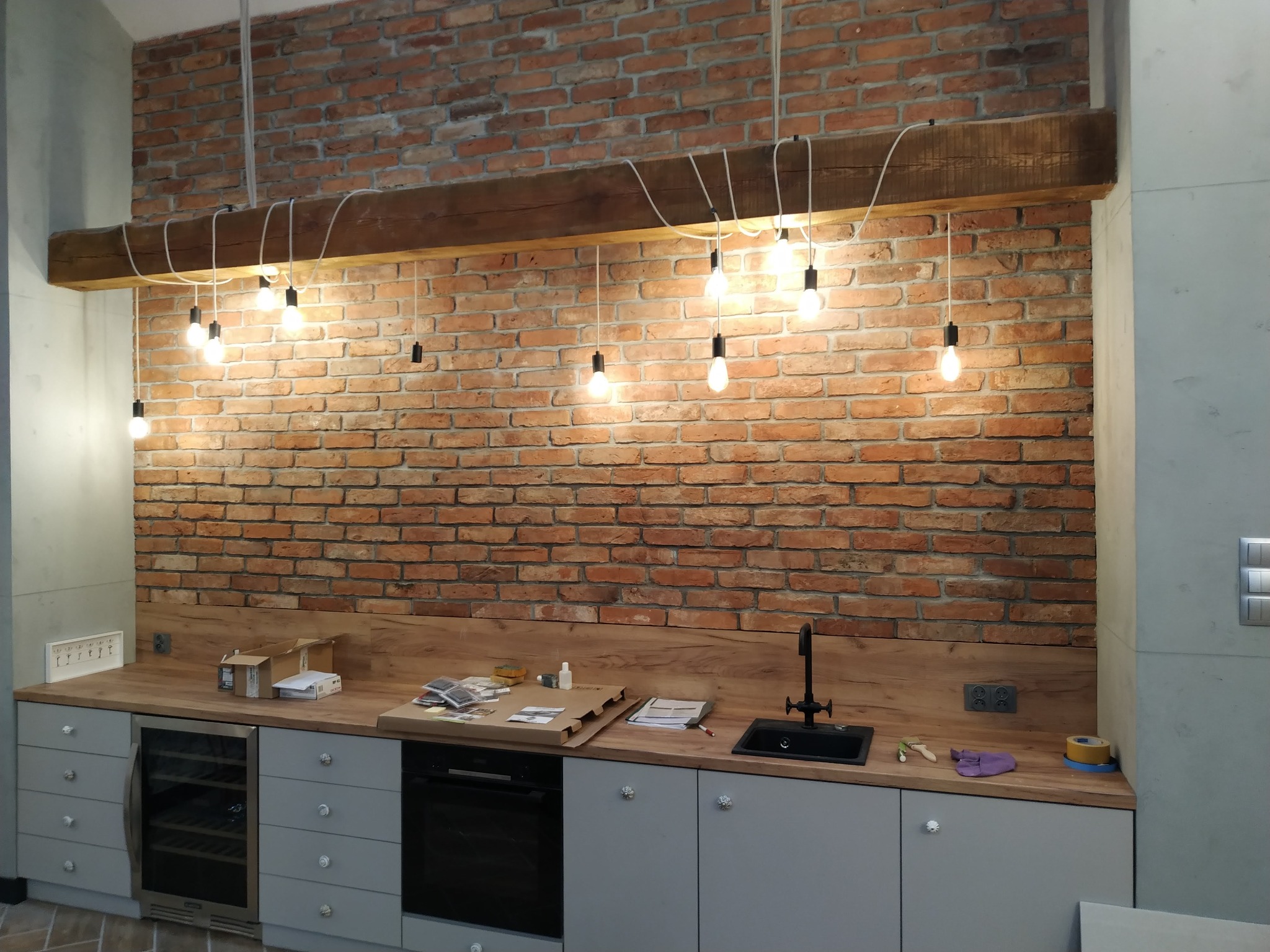 Kuchnia z ceglaną ścianą i wiszącymi żarówkami Edison na drewnianej belce, widoczne gniazdka elektryczne i blat z akcesoriami montażowymi.