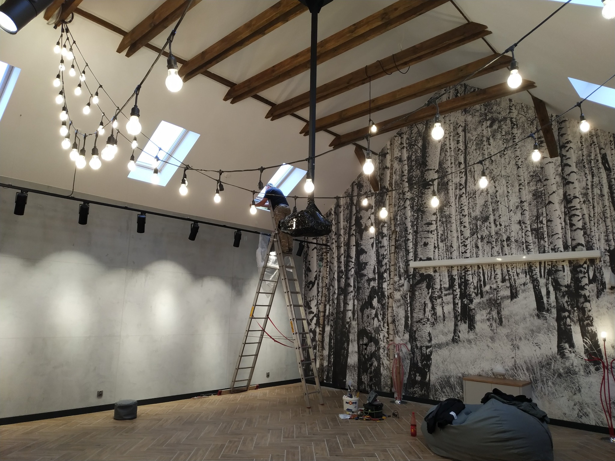 Pracownik na drabinie instaluje girlandę żarówkową w pomieszczeniu z betonową ścianą i fototapetą z motywem brzozowego lasu, widoczne narzędzia i elementy oświetlenia.