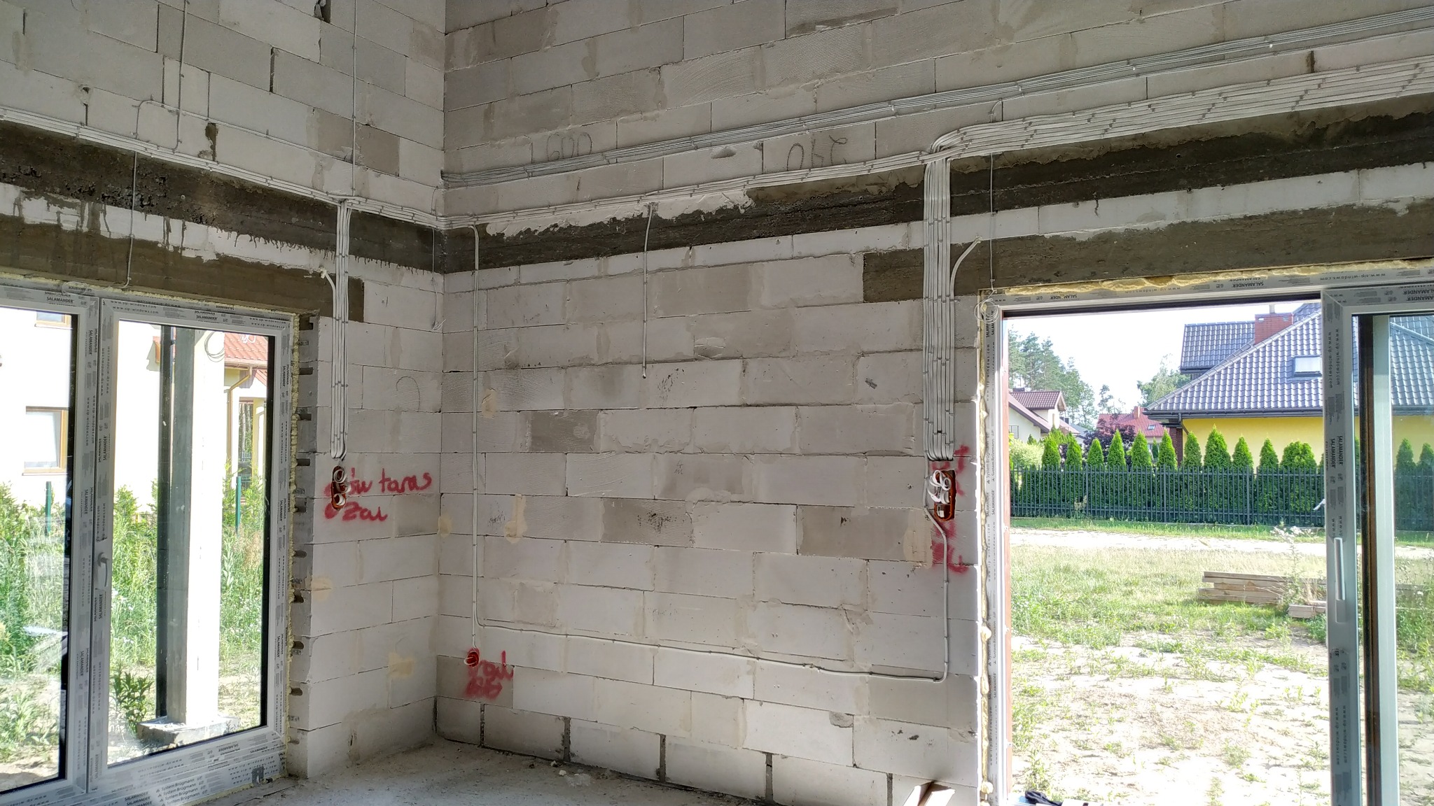 Surowe wnętrze w trakcie budowy z widocznymi przewodami elektrycznymi ułożonymi w korytach na ścianach z betonu komórkowego, czerwone oznaczenia na ścianach, widok przez okno na dom i zieleń.