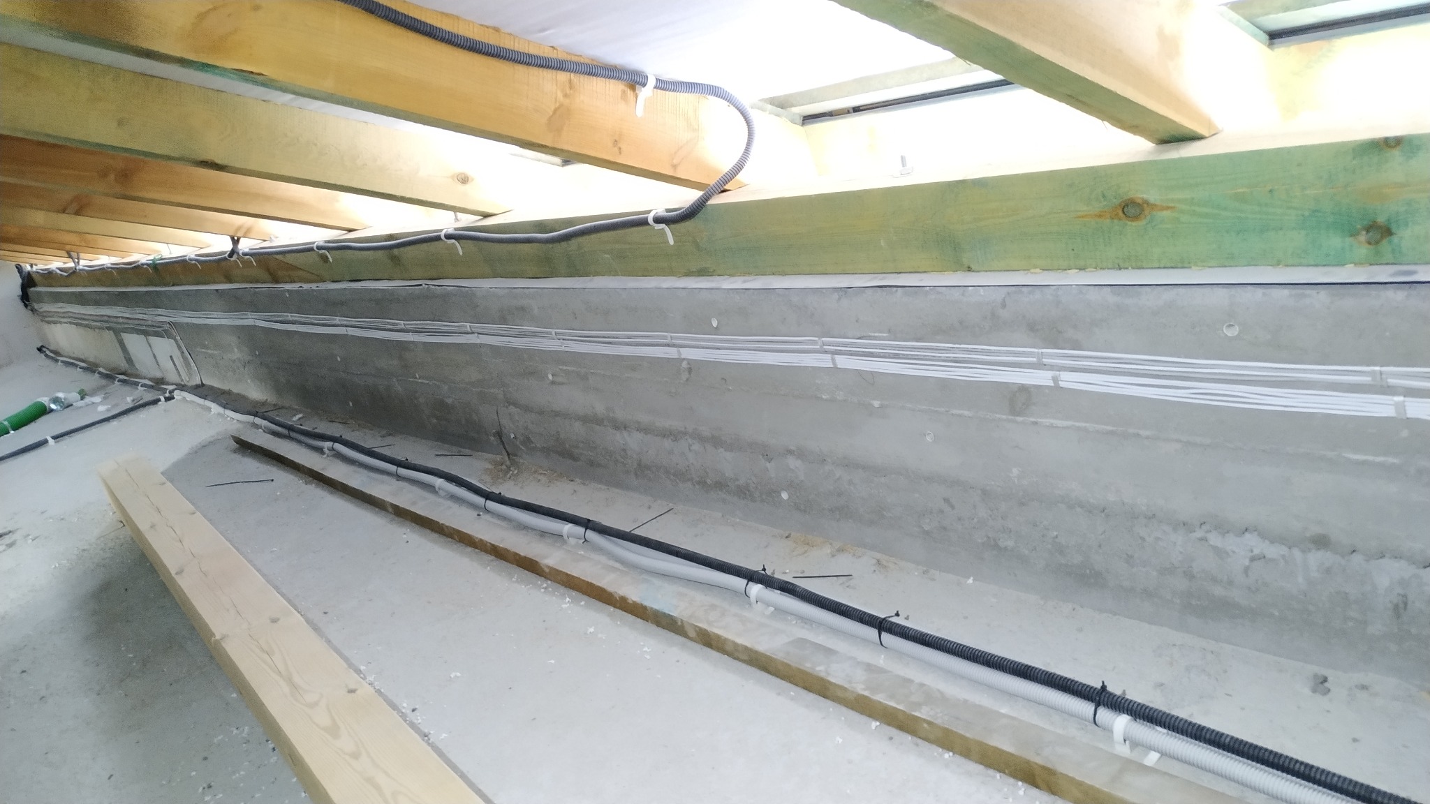 Instalacja elektryczna: Szare kable w peszlach i białe przewody ułożone wzdłuż betonowej ściany pod drewnianą konstrukcją stropu.