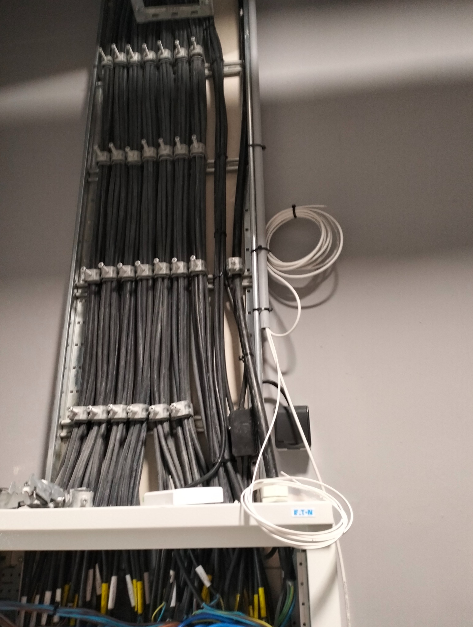 Szara drabinka kablowa z uporządkowanymi przewodami elektrycznymi w budynku. Widoczne kable w peszlach oraz elementy montażowe.