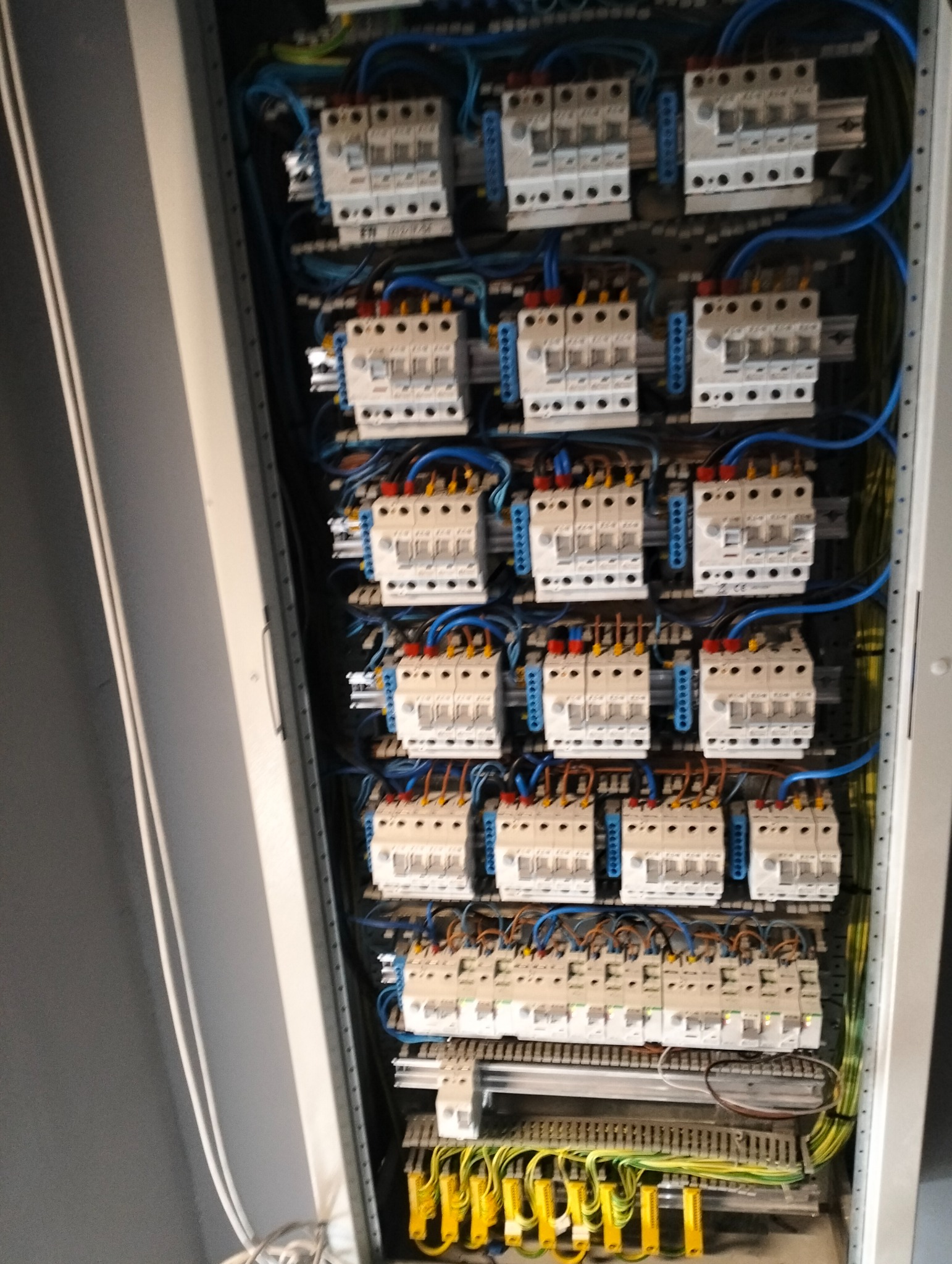 Rozdzielnica elektryczna z licznymi zabezpieczeniami, okablowaniem w kolorach: niebieskim, żółtym i brązowym. Widoczne bezpieczniki i listwy zaciskowe.