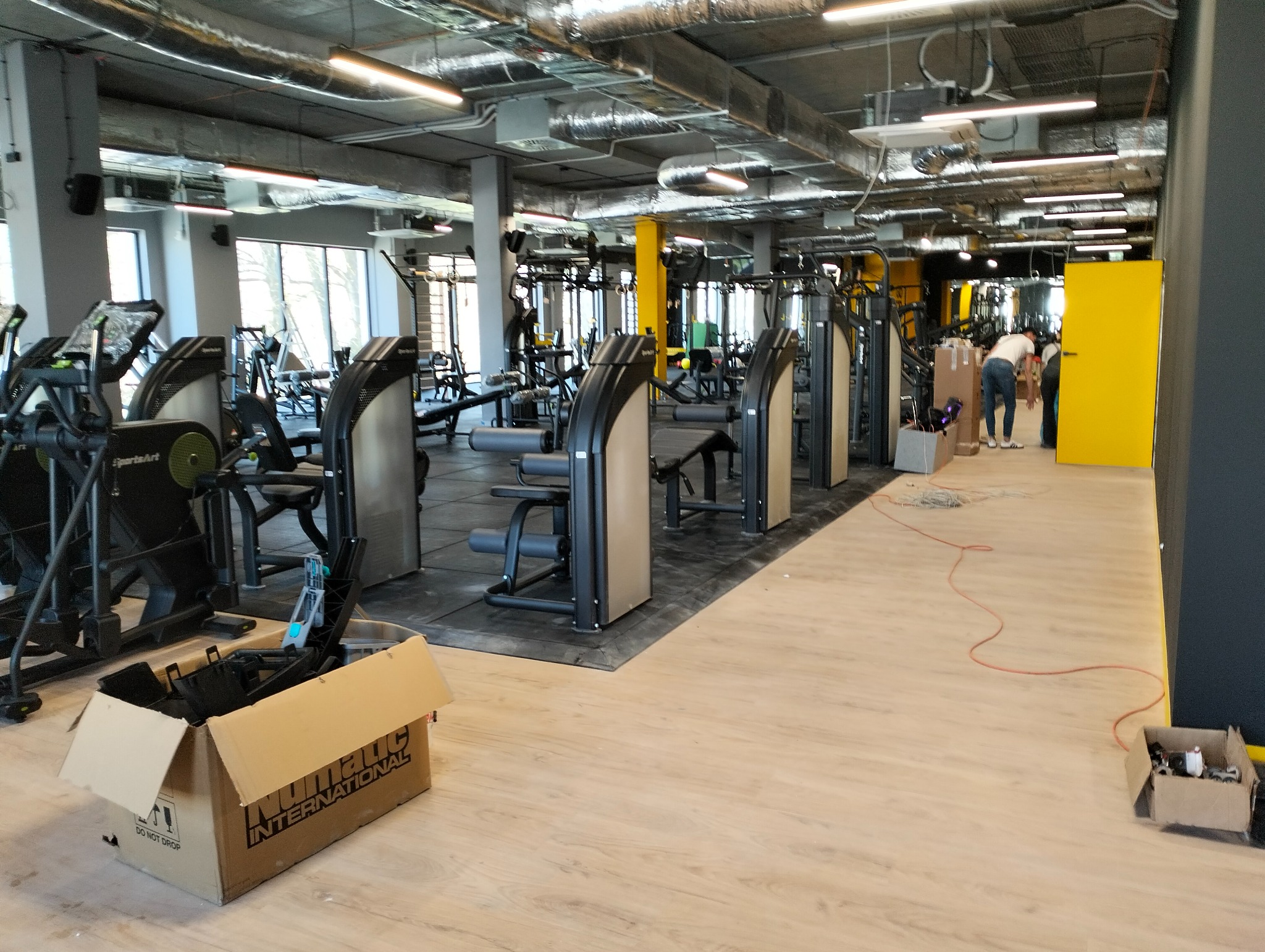 Siłownia w aquaparku po instalacji elektrycznej. Widoczne maszyny do ćwiczeń, kable i skrzynki elektryczne. Oświetlenie sufitowe z elementami instalacji.