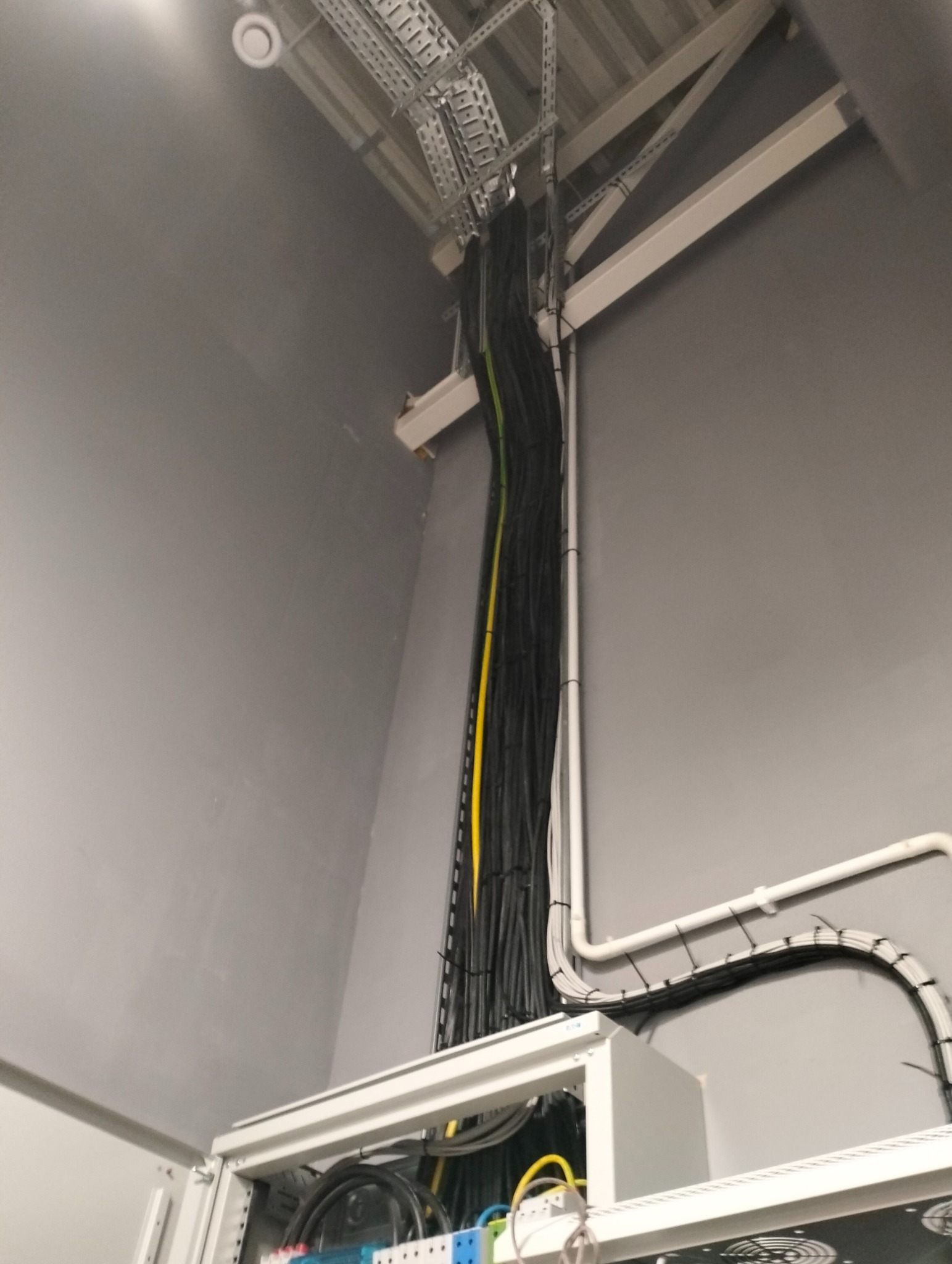 Kompleksowa instalacja elektryczna i teletechniczna w siłowni: widok na prowadnice kablowe, kable w kolorach czarnym, żółtym i zielonym, oraz szafę rack z osprzętem.