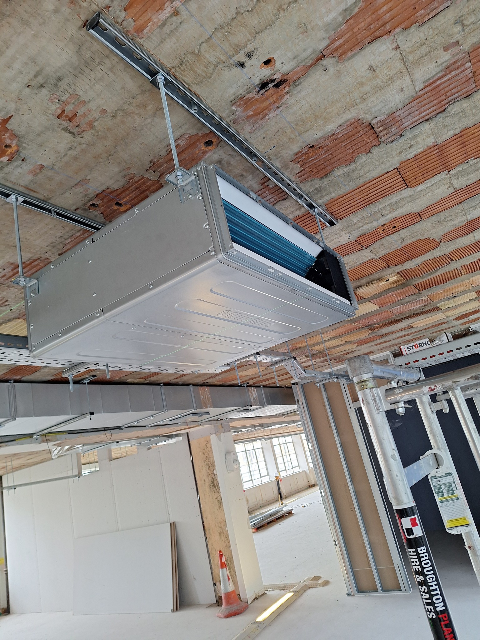 Montaż centrali wentylacyjnej pod sufitem z widoczną konstrukcją nośną i elementami instalacji. Widok z dołu, surowy stan wykończenia wnętrza.