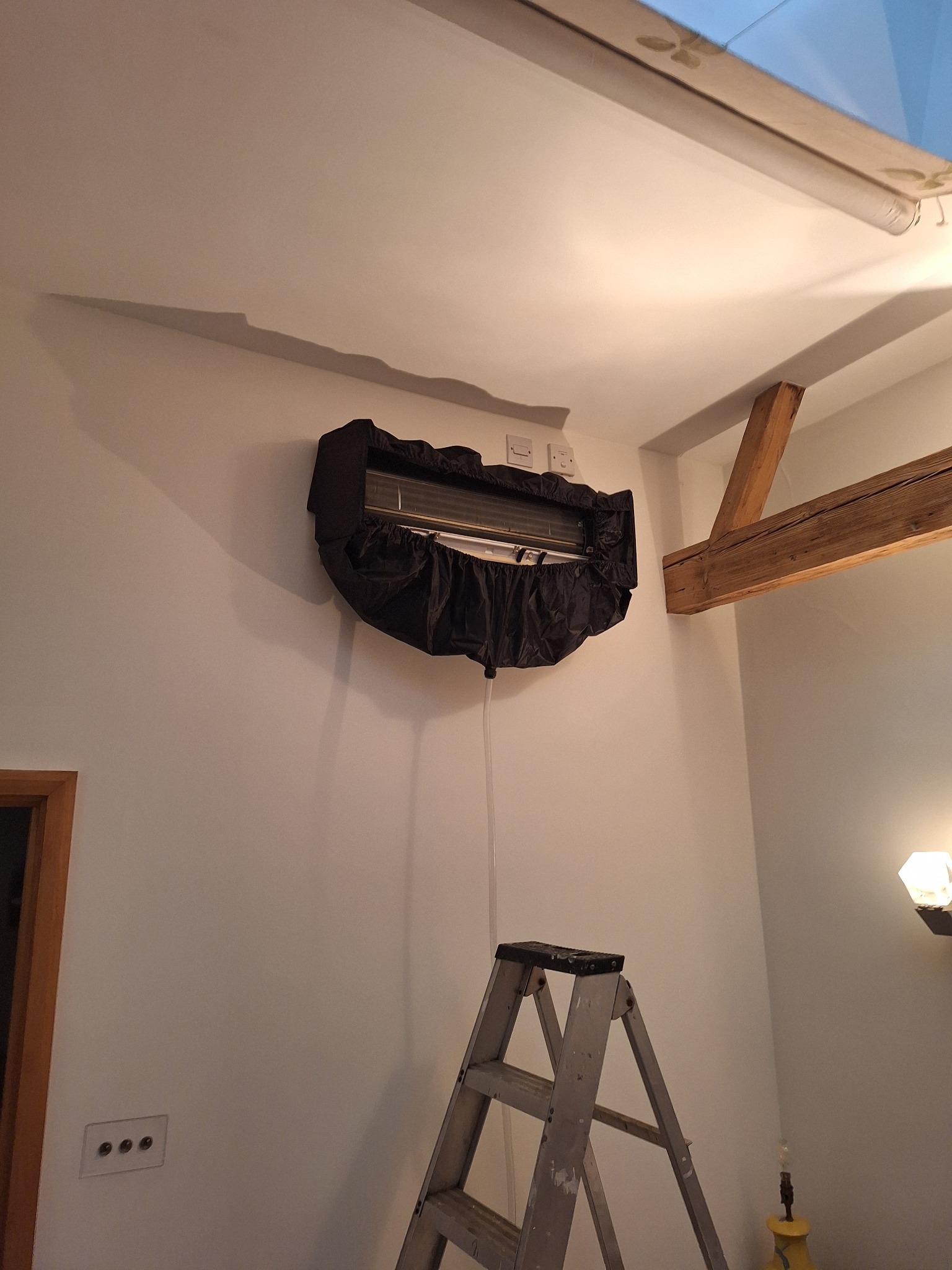 Klimatyzator zabezpieczony czarną osłoną przed czyszczeniem, drabina pod spodem. Przygotowanie do serwisu w pomieszczeniu z drewnianymi elementami.