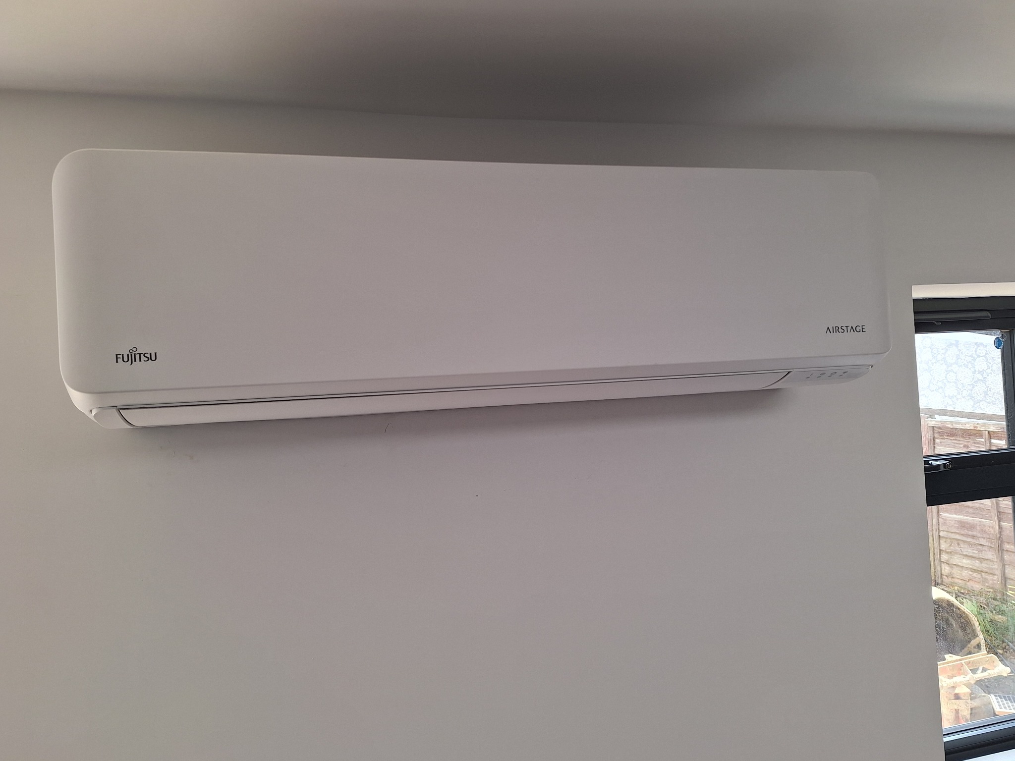 Biała klimatyzacja Fujitsu Airstage zamontowana na ścianie w pomieszczeniu, widok z boku, z oknem w tle.