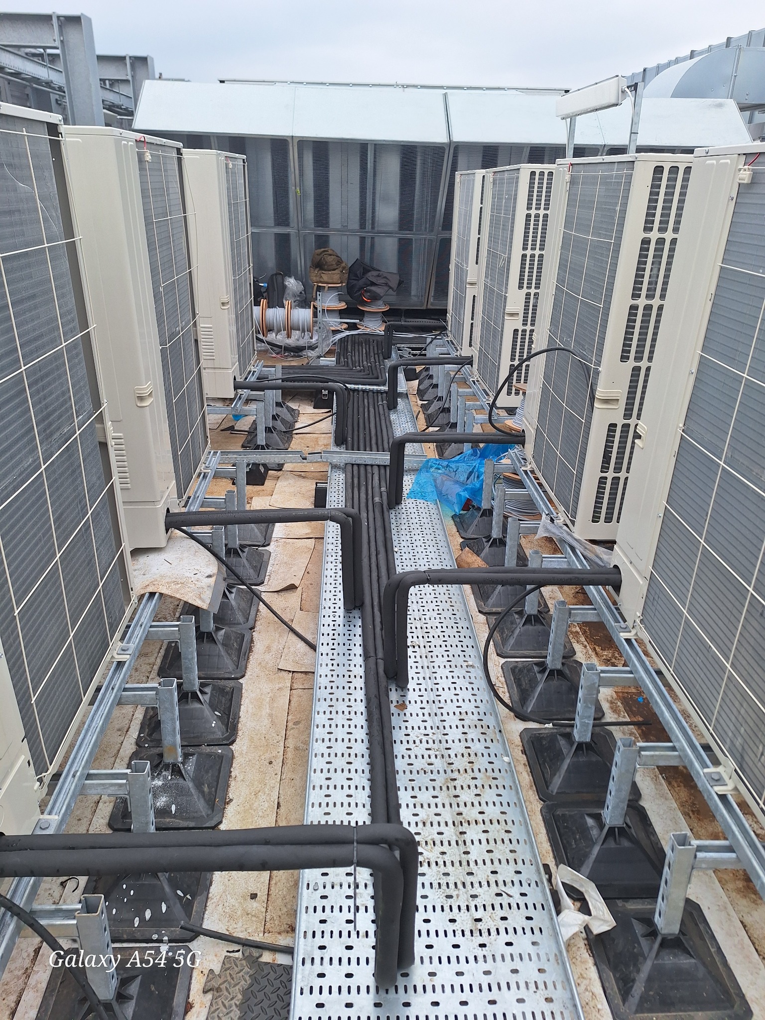 Rząd zewnętrznych jednostek klimatyzacyjnych na dachu budynku, połączonych czarnymi rurami. Widoczne metalowe wsporniki i kable. Ujęcie z perspektywy.