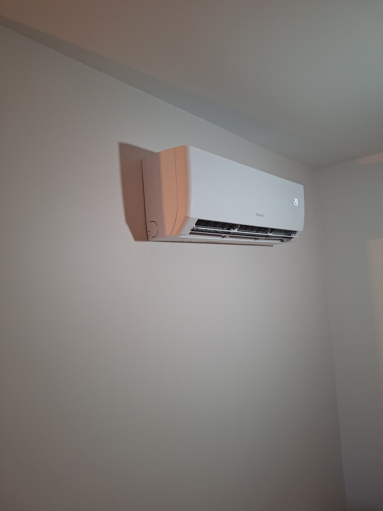 Biały klimatyzator marki Gree zamontowany na jasnej ścianie. Widoczny wyświetlacz z temperaturą 28 stopni. Minimalistyczny design, wnętrze pomieszczenia.